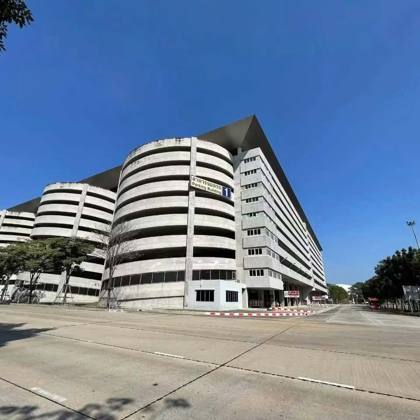 ภาพถ่ายอาคารจอดรถคอนกรีตหลายชั้นทรงโค้งขนาดใหญ่ มีป้าย 'อาคารจอดรถ Parking Building 1' ใต้ท้องฟ้าสีฟ้าสด ถนนด้านหน้าว่างเปล่า
